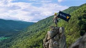 A WVU student carrying a flag that reads "Let's Go." on top of a mountain peak.